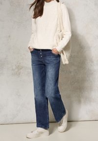 Suéter blanco de punto trenzado, con cuello alto; jeans de mezclilla con botones, de pierna recta, azul medio a oscuro; zapatillas blancas; bolso de mano texturizado.