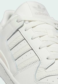 Lähikuva valkoisista Adidas-tenniskenkästä, jossa on kuvioidut raidat, valkoiset nauhat ja Adidas-logo kieleltä.