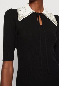 Robe en maille côtelée noire avec un col blanc orné de perles et de paillettes, manches courtes, et un décolleté fendu avec un lien.