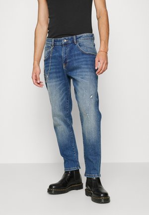 Mann trägt verblasste blaue Jeans mit leichten Gebrauchsspuren, eine Kette, die an einer Gürtelschlaufe befestigt ist, schwarze Chelsea-Stiefel und ein schwarzes ärmelloses Oberteil.