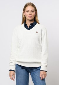Hvit kabelstrikket v-hals genser med ribbestrikkede mansjetter og kant, med en liten brodert logo. Bæres over en mørk skjorte med krage.