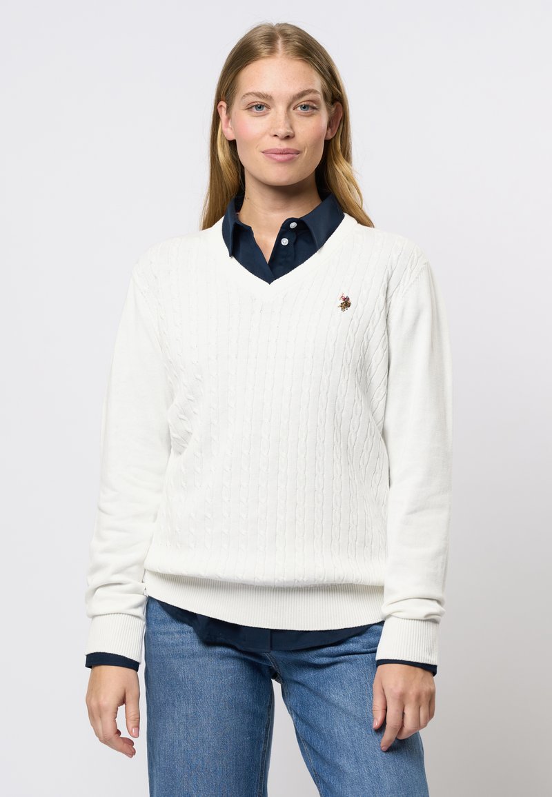 Hvit kabelstrikket v-hals genser med ribbestrikkede mansjetter og kant, med en liten brodert logo. Bæres over en mørk skjorte med krage.