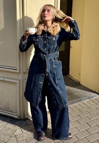 Casaco de ganga com gola de pêlo, botões à frente e cinto de atar; calças largas de ganga; modelo segura uma chávena branca, em pé num pavimento de pedra.