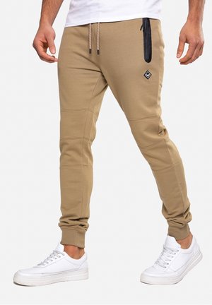 Mann trägt beige Jogginghosen mit seitlicher Reißverschlusstasche und weißen Sneakers, steht vor einem schlichten hellen Hintergrund.