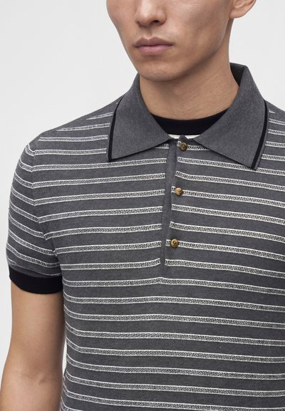 Homme portant un polo rayé gris et noir avec un large col et trois boutons marron sur le devant.