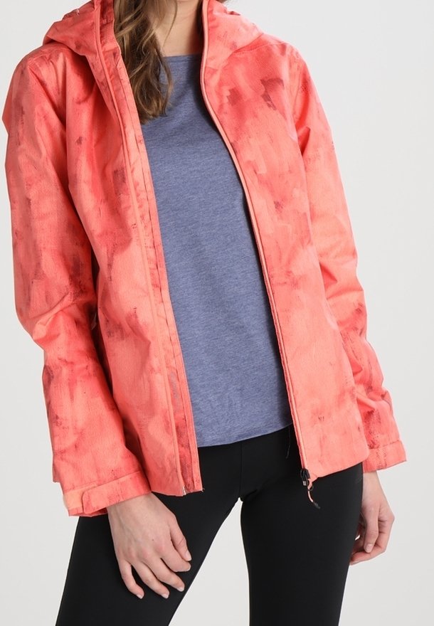 Nainen, joka pukeutunut avoimeen korallikuosiseen hupputakkiin sinisen paidan ja mustien housujen päällä, vaaleaa yksiväristä taustaa vasten.