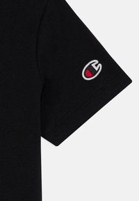 Črna bombažna majica s kratkimi rokavi, ki vsebuje majhen vezen logotip v beli in rdeči barvi. Gladka tekstura z obliko kroga za vrat in čistimi šivi.