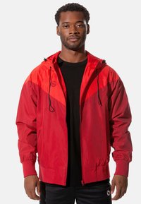 Rote Kapuzenjacke mit einem kontrastierenden dunkler roten oberen Abschnitt, Reißverschluss vorne, elastischen Bündchen und zwei Seitentaschen. Glatte Textur.