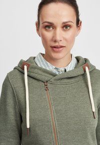 Green zip-up hoodie with a soft texture, featuring a large hood, white drawstrings, and brown hardware accents. Underneath is a blue-striped shirt.