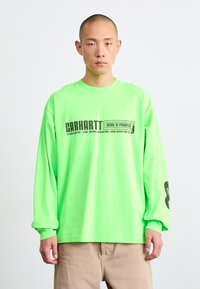 Neon žalios spalvos ilgomis rankov�ėmis marškiniai su juodu "Carhartt Work in Progress" logotipu. Yra gipiūruota apykaklė ir laisvas kirpimas.