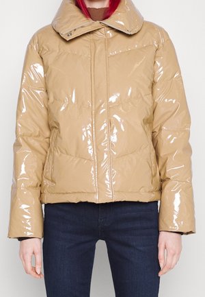 Veste matelassée beige brillante avec grand col portée sur un haut marron et un jean bleu foncé, présentée sur une personne debout, de l'épaule à la hanche.