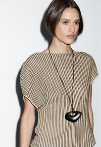 Modella con capelli corti castani che indossa una maglietta a righe beige e un collier nero audace, su uno sfondo bianco.