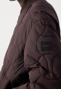 Gros plan d'une personne portant une veste matelassée marron foncé avec un patch logo "BOSS" noir et marron sur la manche supérieure.