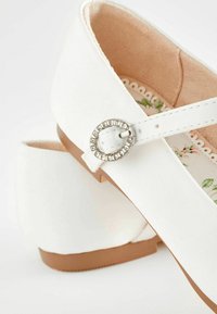 Zapatos de tela blanca con tacón bajo, que presentan un acento de hebilla metálica y forro interior con un diseño floral. Diseño de puntera redondeada.