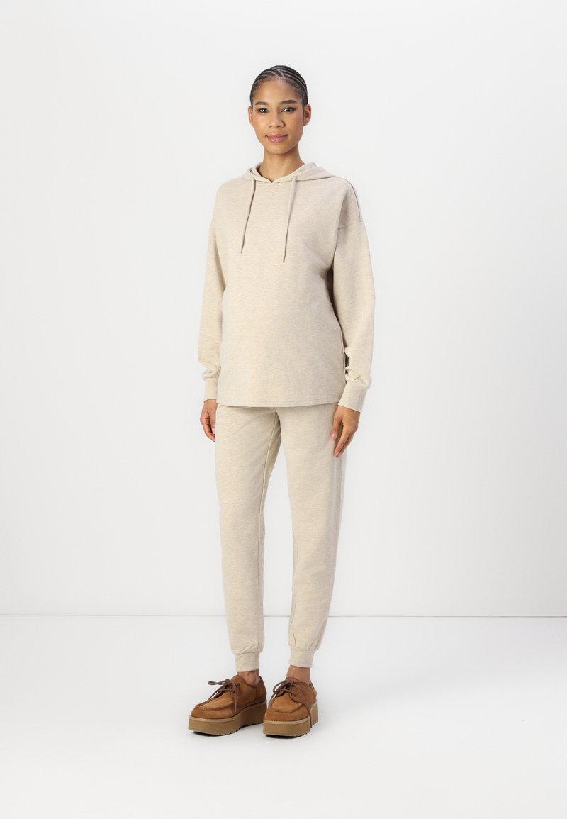 Beige hoodie with drawstrings, paired with matching joggers. Model wearing brown lace-up platform shoes. Smooth texture, relaxed fit.