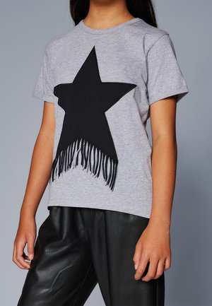 Pige iført en grå t-shirt med korte ærmer med en stor sort stjerne og frynser, kombineret med sorte læderbukser.