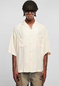 Chemise à manches courtes, de couleur beige clair, avec deux poches poitrine. Texture lisse et devant boutonné, portée avec un jean délavé et de grandes lunettes de soleil noires.