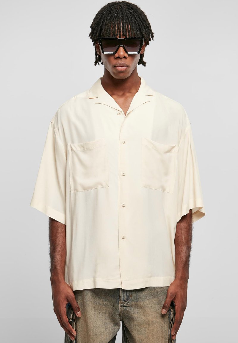 Chemise à manches courtes, de couleur beige clair, avec deux poches poitrine. Texture lisse et devant boutonné, portée avec un jean délavé et de grandes lunettes de soleil noires.