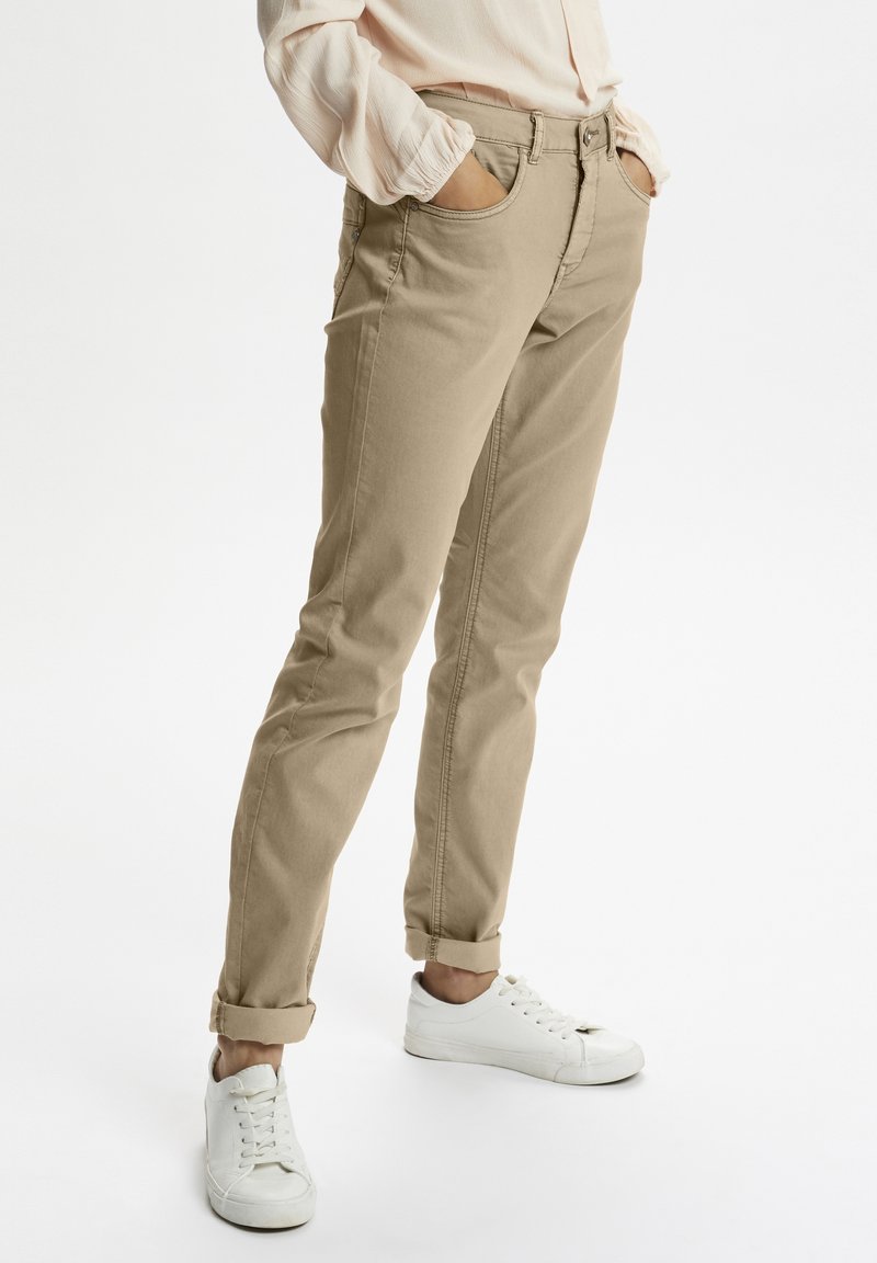 Chinos beige de corte recto y con puños remangados, que incluyen bolsillos frontales y cierre de cremallera, combinados con zapatillas blancas.