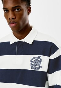 Polo majica prugasta u mornarsko plavoj i bijeloj boji s odgovarajućom kragnom na dugme, s vezanim logotipom na lijevoj strani grudi i teksturiranim materijalom.