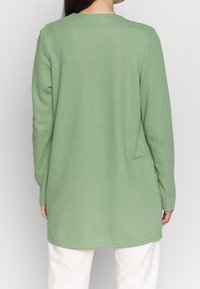 Maglietta a maniche lunghe verde chiaro in tessuto testurizzato, con scollo rotondo e vestibilità ampia, che arriva sotto i fianchi. Parte inferiore bianca visibile.