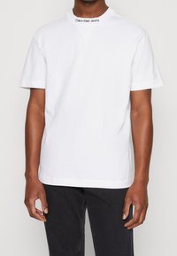 Camiseta de algodón blanca con cuello redondo, mangas cortas y el logo "Calvin Klein Jeans" bordado en el escote. Diseño simple y limpio.