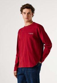 Maglietta rossa a maniche lunghe in cotone con scollo rotondo e scritta bianca sul petto. Abbinate a jeans di denim blu. Texture liscia, vestibilità comoda.