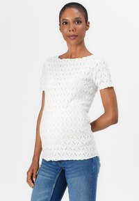 Hvit kortermet topp med et teksturert diamantmønster, rund hals og bølgete kanter. Sammen med blå denimjeans.