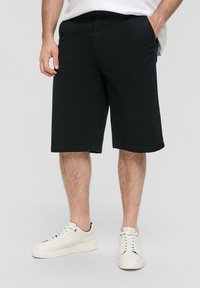 Zwarte knielange shorts met een gladde textuur, voorzien van zijzakken en een rechte snit. Gecombineerd met witte leren sneakers.