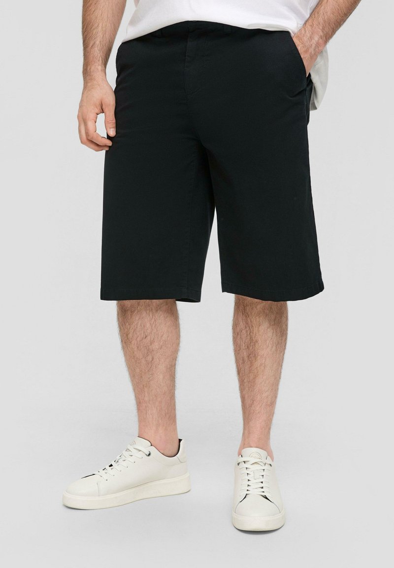 Zwarte knielange shorts met een gladde textuur, voorzien van zijzakken en een rechte snit. Gecombineerd met witte leren sneakers.