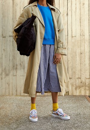 Mujer con un abrigo beige, suéter azul, falda a cuadros en blanco y negro, calcetines amarillos, zapatillas coloridas, llevando una bolsa negra.