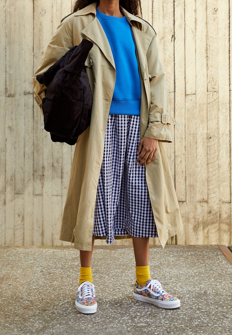 Femme portant un trench-coat beige, un pull bleu, une jupe à carreaux noir et blanc, des chaussettes jaunes, des baskets colorées, tenant un sac noir.