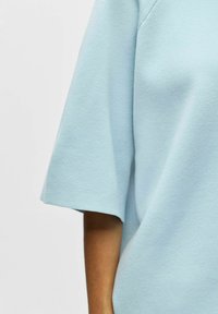 Gros plan d'une personne portant un haut à manches courtes bleu clair, montrant le haut du bras et une partie du torse sur un fond uni.