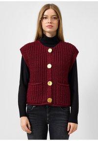 Burgundijas adīta veste ar teksturētu apdari, īsām piedurknēm, pieciem zelta pogām un divām priekšējām kabatām, valkāta pāri melnam turtleneck. 