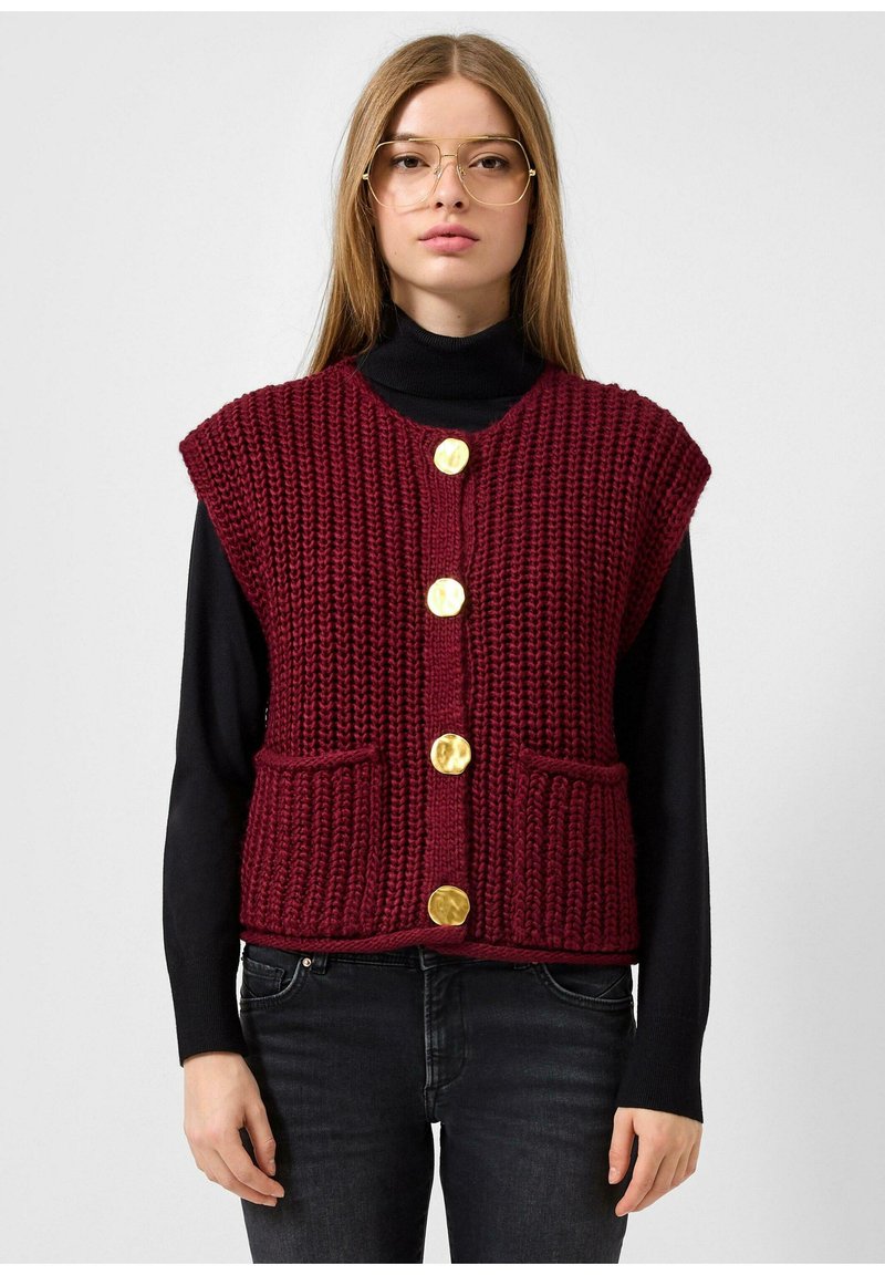 Burgundijas adīta veste ar teksturētu apdari, īsām piedurknēm, pieciem zelta pogām un divām priekšējām kabatām, valkāta pāri melnam turtleneck. 