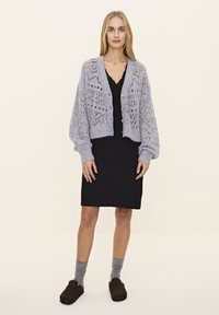 Hellgraue Strickjacke mit aufwendigen Spitzenmustern, mit Knopfverschluss, getragen über einem schwarzen Kleid und grauen Socken, kombiniert mit dunklen Schuhen.