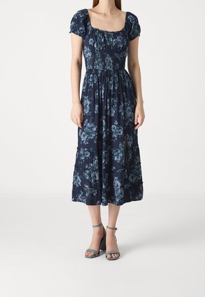 Woman wearing a navy blue floral midi dress with short puff sleeves, smocked waist, front slit, and ankle-strap block heel sandals.