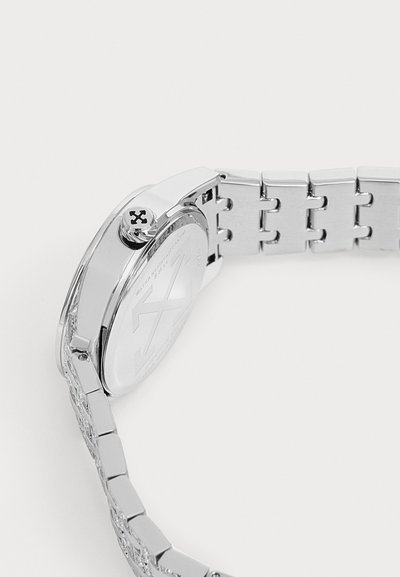 Montre-bracelet en métal argenté avec bracelet à maillons et emblème en croix sur la couronne, vue de côté et de dos sur fond blanc.
