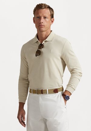 Beige Langarm-Polohemd mit kleinem weißen Logo. Kombiniert mit weißen Hosen und einem geflochtenen, beigen Gürtel mit braunen Details. Dunkle Sonnenbrille am Kragen.