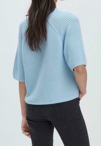 Femme aux longs cheveux foncés portant un pull en maille bleu clair à manches courtes et un jean noir, debout, une main dans la poche, dos tourné vers la caméra.