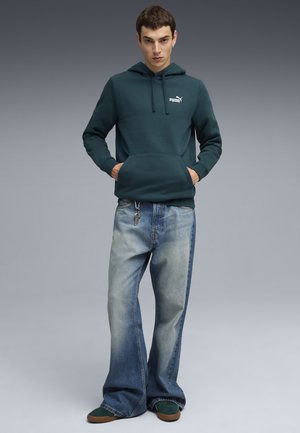 Diep teal hoodie met een voorzak en een wit Puma-logo, gecombineerd met lichtblauwe flared jeans met een vervaagd ontwerp en sleutelhangerdetail.