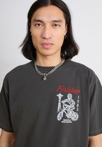 T-shirt court à manches courtes gris foncé avec un imprimé graphique d'un rider BMX et le texte "Finisher 1983 Seattle BMX Racing", accompagné d'un collier en chaîne argenté.