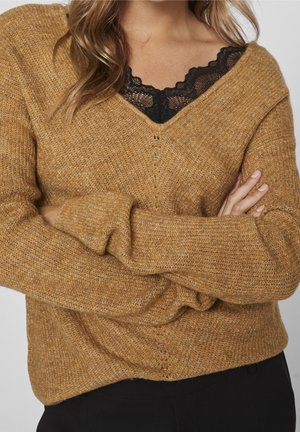 Femme portant un pull marron texturé à col en V par-dessus un haut en dentelle noire, les bras croisés, avec des cheveux châtain clair tombant sur ses épaules.