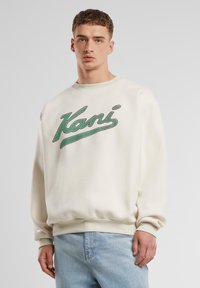 Off-white sweatshirt med grønt "Kani" logo. Har ribbede manchetter og kant, afslappet pasform og en blød tekstur. Bæres med lyseblå jeans.