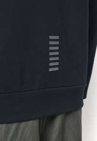 Svart sweatshirt med texturerad yta, som har en vertikal rad av sju reflekterande ränder på nedre vänstra sidan.