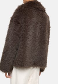 Brązowy płaszcz z sztucznego futra o miękkiej, teksturowanej powierzchni. Posiada szeroki kołnierz i luźny krój, w zestawieniu z jasnymi spodniami.