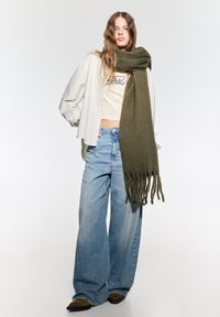 Écharpe vert olive avec des extrémités frangées, portée sur un top court beige et une veste oversized rayée blanche, associée à un pantalon en jeans large bleu clair.
