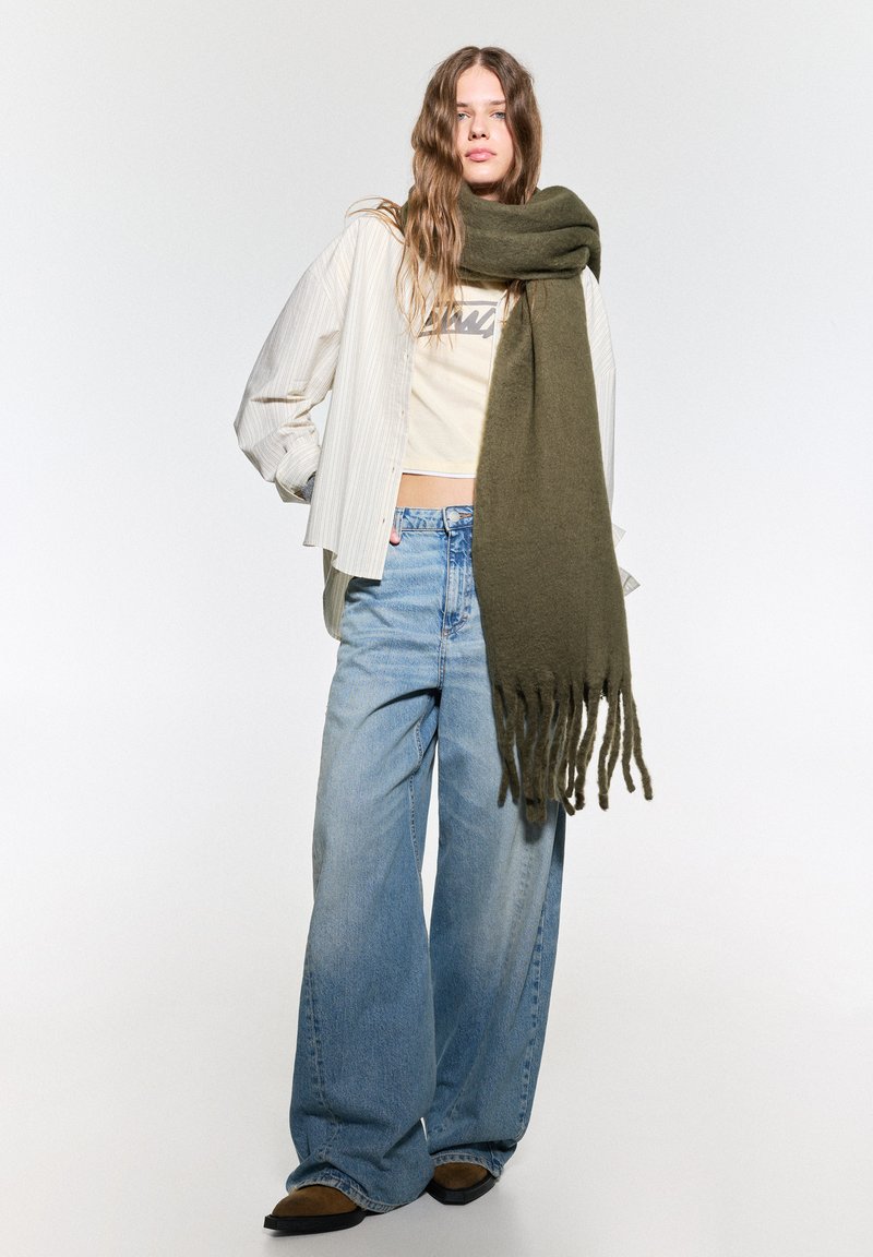 Écharpe vert olive avec des extrémités frangées, portée sur un top court beige et une veste oversized rayée blanche, associée à un pantalon en jeans large bleu clair.