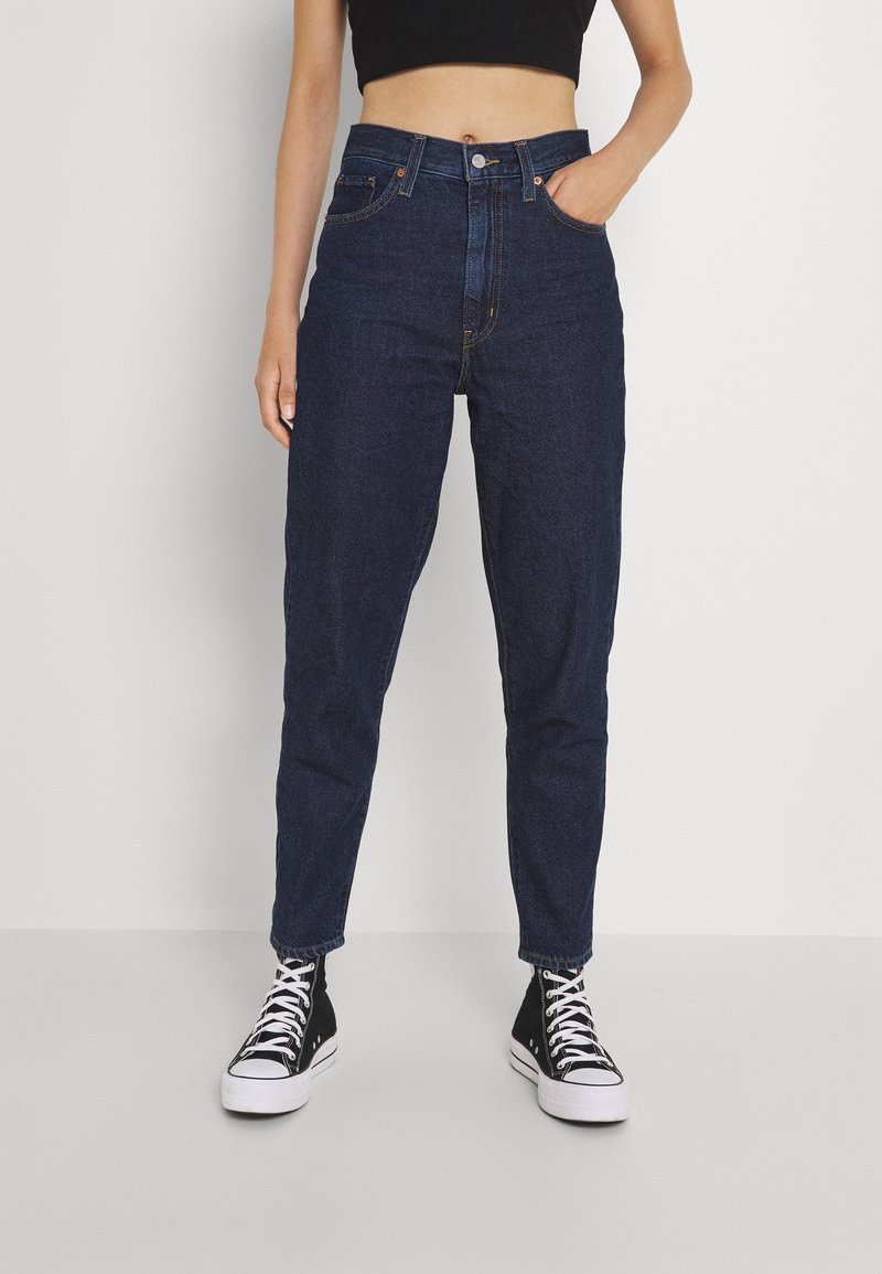 Jeans de mezclilla oscuros de cintura alta con un diseño de pierna recta; cuenta con bolsillos frontales y un cierre de cremallera. Combinados con zapatillas negras de caña alta.