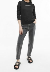 Sudadera negra con el logotipo blanco de "Calvin Klein Jeans", combinada con jeans de mezclilla grises y sandalias negras con un diseño de hebilla.